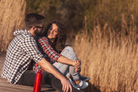 Téléchargez les photos : Jeune couple amoureux assis sur les quais du lac, profitant d'une belle journée d'automne ensoleillée dans la nature - en image libre de droit