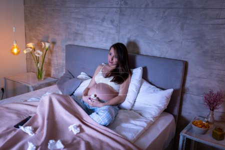 Téléchargez les photos : Femme enceinte portant un pyjama assis sur le lit et pleurant en regardant un film triste à la télévision - en image libre de droit