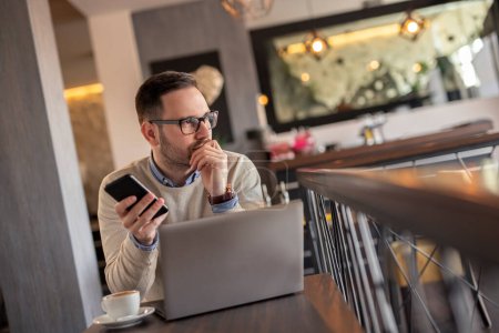 Foto de Freelancer masculino trabajando remotamente desde un restaurante, sosteniendo un teléfono inteligente y utilizando una computadora portátil, seria y pensativa - Imagen libre de derechos
