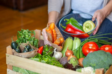 Photo for Detail of woman holding a bowl and picking out fresh organic vegetables for lunch from a food delivery wooden crate - Royalty Free Image