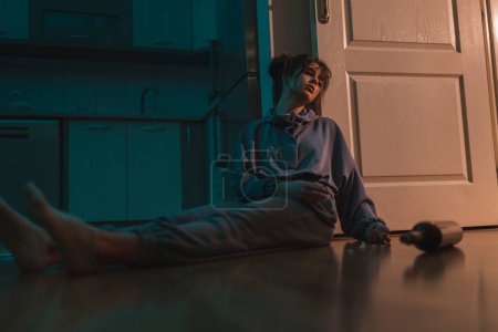 Téléchargez les photos : Jeune femme déprimée assise par terre dans l'obscurité, bouleversée et désespérée, ivre et surdosée de pilules - en image libre de droit