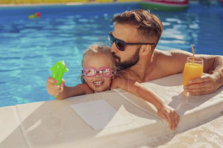 Téléchargez les photos : Belle petite fille et son père s'amusent à la piscine par une chaude journée d'été ensoleillée, le père étreignant et embrassant sa fille pendant qu'elle joue avec un pistolet à éjaculation féminine - en image libre de droit