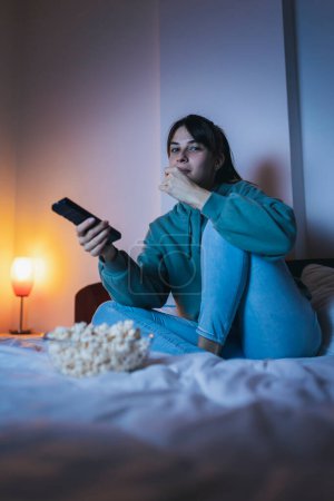 Téléchargez les photos : Belle jeune femme en pyjama assise au lit, mangeant du pop-corn et regardant la télévision, se relaxant à la maison tard dans la nuit - en image libre de droit