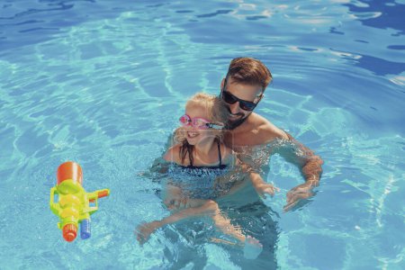 Téléchargez les photos : Vue en angle élevé de la belle petite fille et son père s'amusant dans la piscine par une chaude journée d'été ensoleillée, père enseignant à sa fille à nager - en image libre de droit
