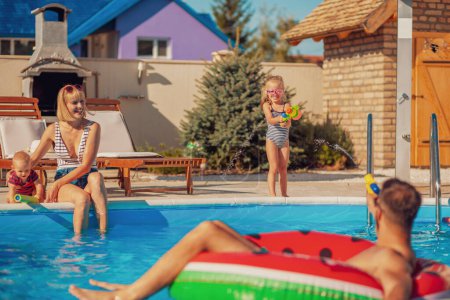 Téléchargez les photos : Les parents joyeux s'amusent à jouer avec leurs enfants avec des pistolets à éjaculation près de la piscine éclaboussant l'eau les uns sur les autres, profitant des vacances d'été - en image libre de droit