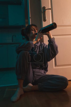 Photo for Depressed drunk woman sitting on the floor in the dark with smeared makeup, drinking wine from a bottle and crying - Royalty Free Image