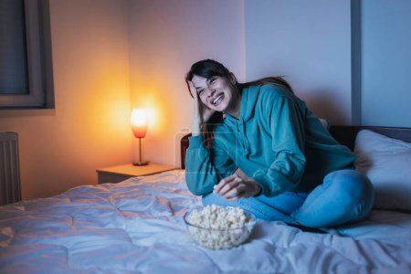 Téléchargez les photos : Belle jeune femme portant un pyjama assis au lit, mangeant du pop-corn et riant tout en regardant un film drôle à la télévision, se détendre à la maison tard dans la nuit - en image libre de droit