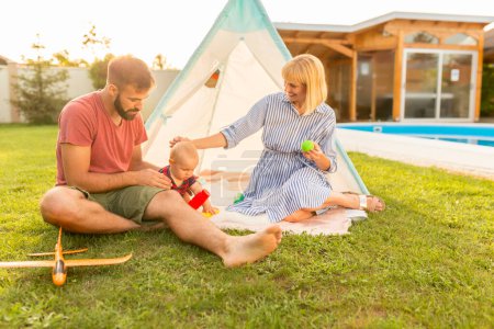 Téléchargez les photos : De jeunes parents heureux s'amusent à camper et à jouer avec leur petit garçon au bord de la piscine dans la cour arrière de leur maison - en image libre de droit