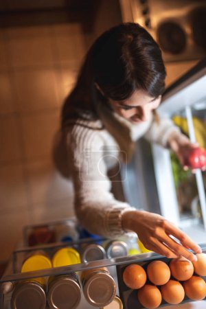 Téléchargez les photos : Vue de dessus d'une belle jeune femme prenant des produits alimentaires d'un réfrigérateur ouvert. Focus sur les œufs - en image libre de droit