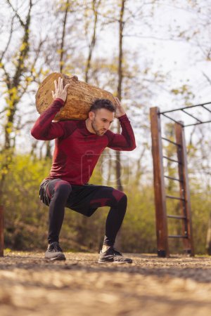 Téléchargez les photos : Vue à angle bas d'un homme musclé actif soulevant un tronc d'arbre tout en faisant des squats, s'exerçant à l'extérieur dans un parc d'entraînement de rue - en image libre de droit