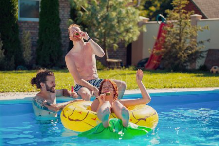 Gruppe junger Freunde vergnügt sich bei einer sommerlichen Poolparty, verbringt einen sonnigen Sommertag im Freien und isst Wassermelonen-Eis am Pool