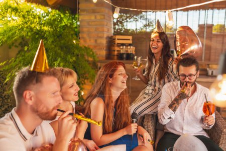 Photo for Group of cheerful young friends having fun at a birthday party, singing birthday song, blowing party whistles and drinking cocktails - Royalty Free Image