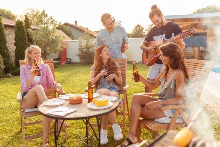 Foto de Grupo de amigos divirtiéndose tocando la guitarra y cantando en la fiesta de barbacoa del patio trasero, bebiendo cerveza y relajándose al aire libre en un día soleado de verano - Imagen libre de derechos