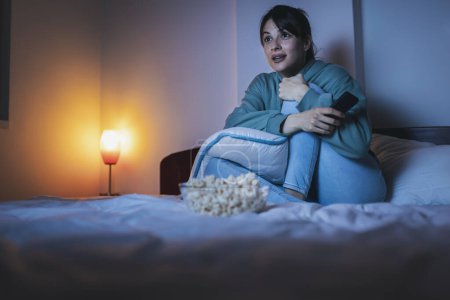Téléchargez les photos : Belle jeune femme en pyjama assise au lit la nuit, mangeant du pop-corn et regardant un film à la télé - en image libre de droit