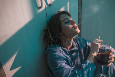 Photo for Young woman sitting on the floor in the dark, drinking wine and smoking cigarette, depressed and alone - Royalty Free Image