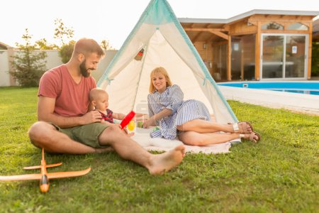 Photo for Young parents having fun camping and playing with their little baby boy by the swimming pool in the backyard of their house - Royalty Free Image
