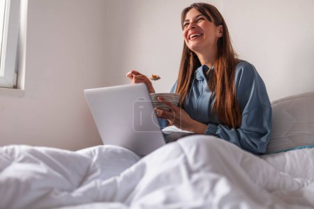 Foto de Mujer joven usando pijamas sentada en la cama por la mañana, desayunando en la cama, comiendo cereales y usando computadora portátil - Imagen libre de derechos