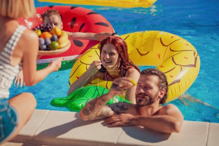 Grupo de jóvenes amigos divirtiéndose, relajándose y refrescándose en la piscina en un caluroso día de verano, comiendo fruta y flotando en colchones de aire