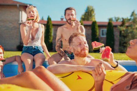 Foto de Grupo de jóvenes amigos divirtiéndose en la fiesta de verano en la piscina, pasando el soleado día de verano al aire libre y comiendo paletas de sandía junto a la piscina - Imagen libre de derechos