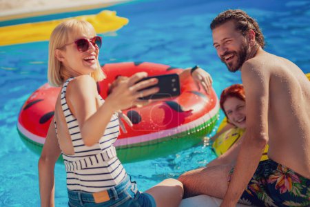 Téléchargez les photos : Deux beaux jeunes couples s'amusent à la piscine par une journée ensoleillée d'été, prenant un selfie à l'aide d'un téléphone intelligent, se détendre et bronzer au bord de la piscine - en image libre de droit