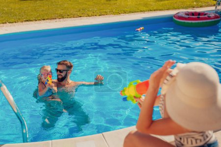 Foto de Los padres jóvenes se divierten jugando con su hija en la piscina, salpicando agua con pistolas de agua, disfrutando de un caluroso día soleado de verano al aire libre y relajándose mientras están en vacaciones de verano. - Imagen libre de derechos