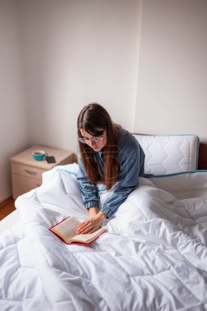 Téléchargez les photos : Belle jeune femme coûteuse portant un pyjama assis au lit le matin, lisant un livre et se relaxant à la maison - en image libre de droit