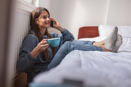 Téléchargez les photos : Belle jeune femme portant un pyjama assis par terre près du lit dans la chambre, buvant du café et ayant une conversation téléphonique - en image libre de droit