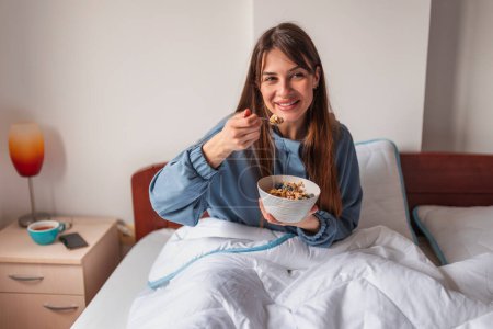 Foto de Hermosa joven con pijamas sentada en la cama por la mañana, tomando cereales con bayas frescas para el desayuno y disfrutando del tiempo libre en casa - Imagen libre de derechos