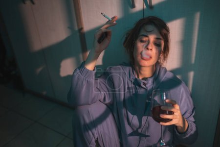 Photo for Depressed young woman sitting on the floor in the dark, drinking wine and smoking cigarette, upset and desperate - Royalty Free Image