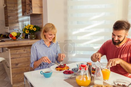 Photo for Beautiful young couple in love sitting at dinning room table, having waffles, berries, orange juice and coffee for breakfast, spending weekend morning together at home - Royalty Free Image