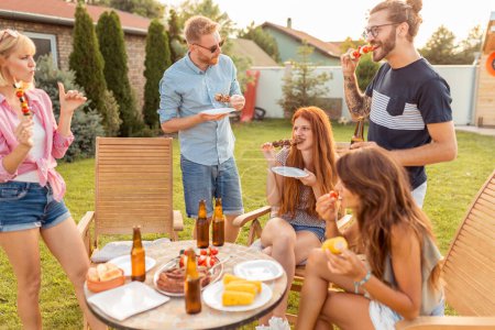 Téléchargez les photos : Groupe de jeunes amis joyeux réunis autour de la table, manger de la viande grillée, boire de la bière et s'amuser à une fête barbecue arrière-cour - en image libre de droit