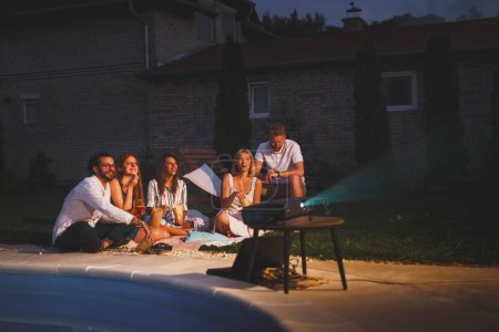 Foto de Grupo de alegres jóvenes amigos divirtiéndose comiendo palomitas de maíz, bebiendo cócteles y viendo una película en un cine al aire libre en el patio trasero de una casa - Imagen libre de derechos