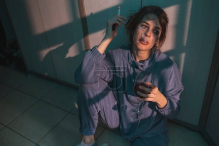 Photo for Depressed young woman sitting on the floor in the dark, drinking wine and smoking cigarette, upset and desperate - Royalty Free Image