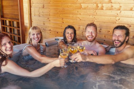 Téléchargez les photos : Groupe de jeunes amis joyeux se détendre dans un centre de spa hôtel spa bain à remous, faire un toast lever des verres de vin et s'amuser pendant les vacances - en image libre de droit