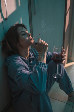 Foto de Mujer joven deprimida sentada en el suelo en la oscuridad, bebiendo vino y fumando cigarrillo, molesta y desesperada - Imagen libre de derechos