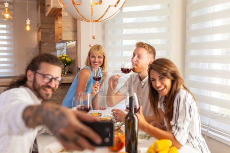 Foto de Grupo de alegres jóvenes amigos divirtiéndose bebiendo vino y tomando selfies mientras cenan juntos en casa - Imagen libre de derechos
