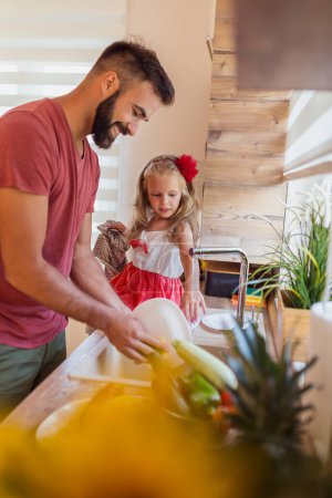 Foto de Hermosa niña sentada en el mostrador de la cocina, divirtiéndose lavando los platos con su padre en la cocina después del almuerzo - Imagen libre de derechos
