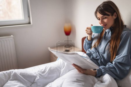 Téléchargez les photos : Belle jeune femme en pyjama se relaxant au lit après le réveil, lisant le journal et buvant son café du matin - en image libre de droit