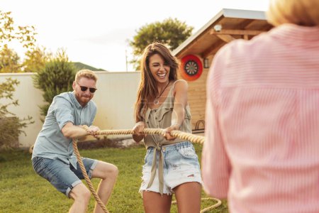 Téléchargez les photos : Groupe de jeunes amis s'amusant tout en participant à une compétition de tire-corde lors d'une fête d'été en plein air ; les gens jouent au remorqueur de guerre - en image libre de droit