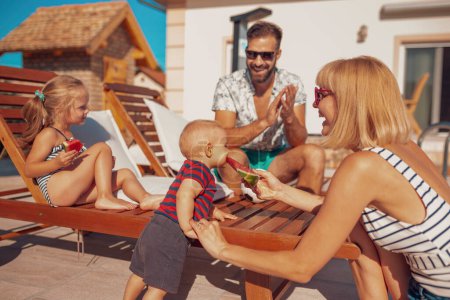 Photo for Parents having fun spending hot sunny summer day by the swimming pool with their children, mother feeding baby boy with a slice of watermelon - Royalty Free Image
