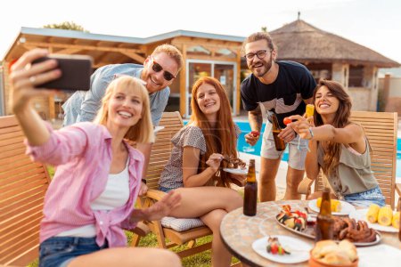 Téléchargez les photos : Groupe d'amis ayant une fête barbecue au bord de la piscine arrière, s'amusant à prendre des selfies tout rassemblés autour de la table - en image libre de droit
