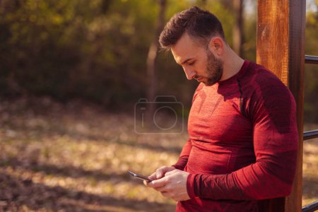 Téléchargez les photos : Jeune homme actif utilisant un téléphone intelligent tout en prenant une pause d'entraînement, en tapant un message texte - en image libre de droit