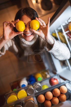 Foto de Vista superior de una hermosa mujer joven de pie junto a una puerta abierta del refrigerador en la cocina por la noche, sonriendo y sosteniendo dos limones sobre los ojos - Imagen libre de derechos