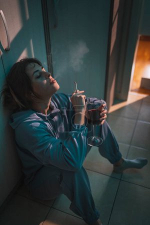 Photo for Young woman sitting on the floor in the dark, drinking wine and smoking cigarette, depressed and alone - Royalty Free Image
