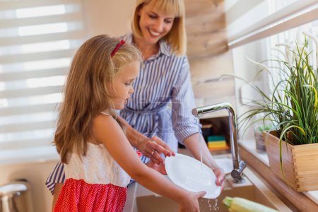 Photo for Beautiful young mother having fun doing the washing up after lunch in the kitchen with her little daughter - Royalty Free Image