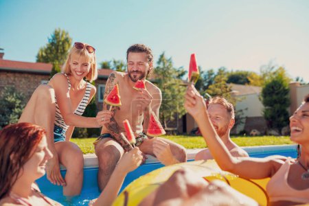 Gruppe junger Freunde, die sich auf der sommerlichen Poolparty amüsieren, am Pool sitzen und Wassermelonen-Eis essen, sonnige Sommertage im Freien verbringen