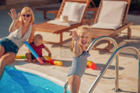 Téléchargez les photos : Belle jeune mère s'amuse à jouer avec ses enfants au bord de la piscine, profitant d'une chaude journée d'été ensoleillée en plein air, mère et fille saluant vers la caméra - en image libre de droit
