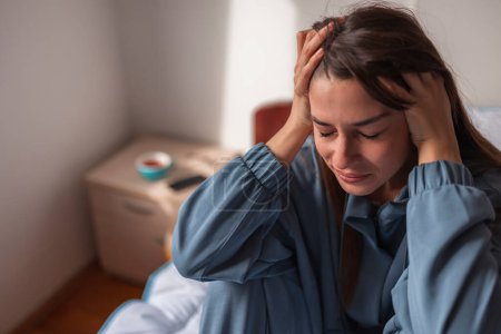 Foto de Mujer joven desesperada usando pijamas sentada en la cama por la mañana, sosteniendo la cabeza en las manos, llorando - Imagen libre de derechos