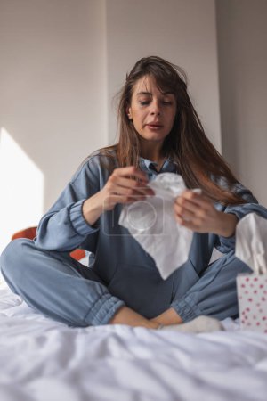 Photo for Depressed young woman wearing pajamas, sitting in bed in the morning, wiping tears and nose using paper tissue while crying - Royalty Free Image