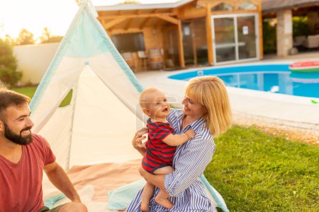 Foto de Padres jóvenes felices divirtiéndose acampando y jugando con su pequeño bebé junto a la piscina en el patio trasero de su casa, madre sosteniendo a su hijo - Imagen libre de derechos